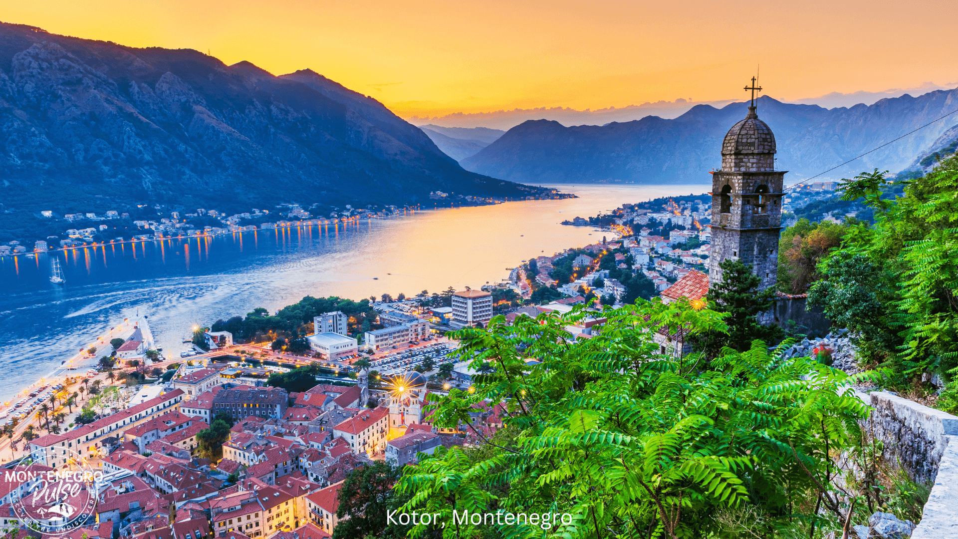 Sunset view of Kotor Bay with red rooftops, the bell tower, and mountains in the distance.