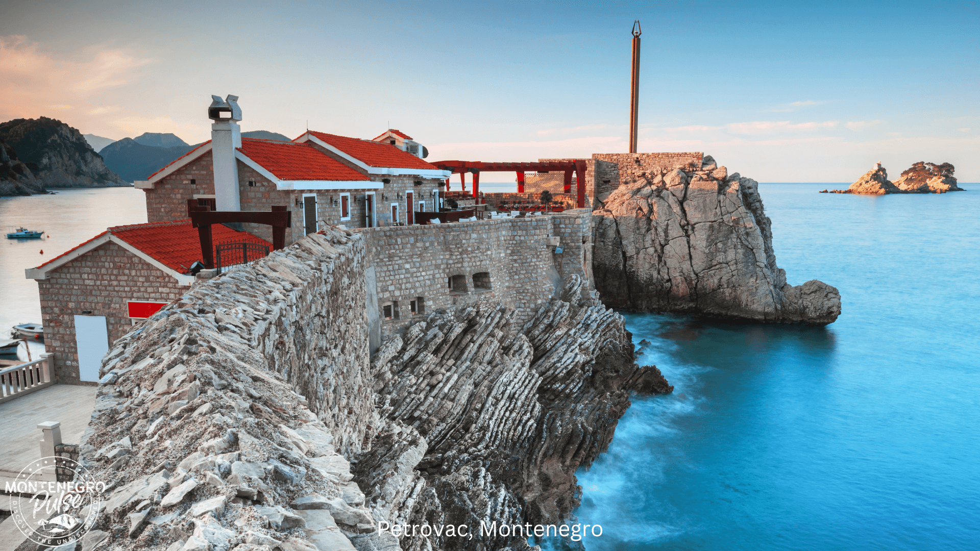 Rocky coastline of Petrovac, Montenegro, with historical buildings and clear blue water at sunset.