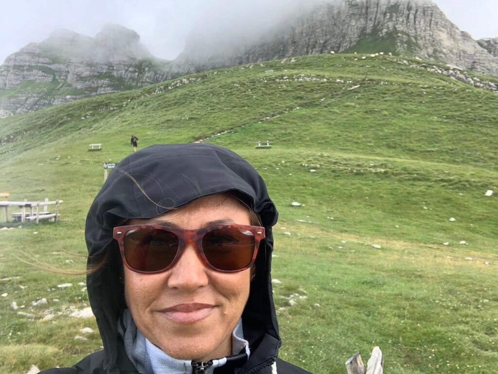 Me wearing sunglasses and a hooded jacket taking a selfie with a scenic mountainous backdrop in Durmitor National Park on a cloudy day; green slopes and rocky outcrops are partially shrouded in mist.